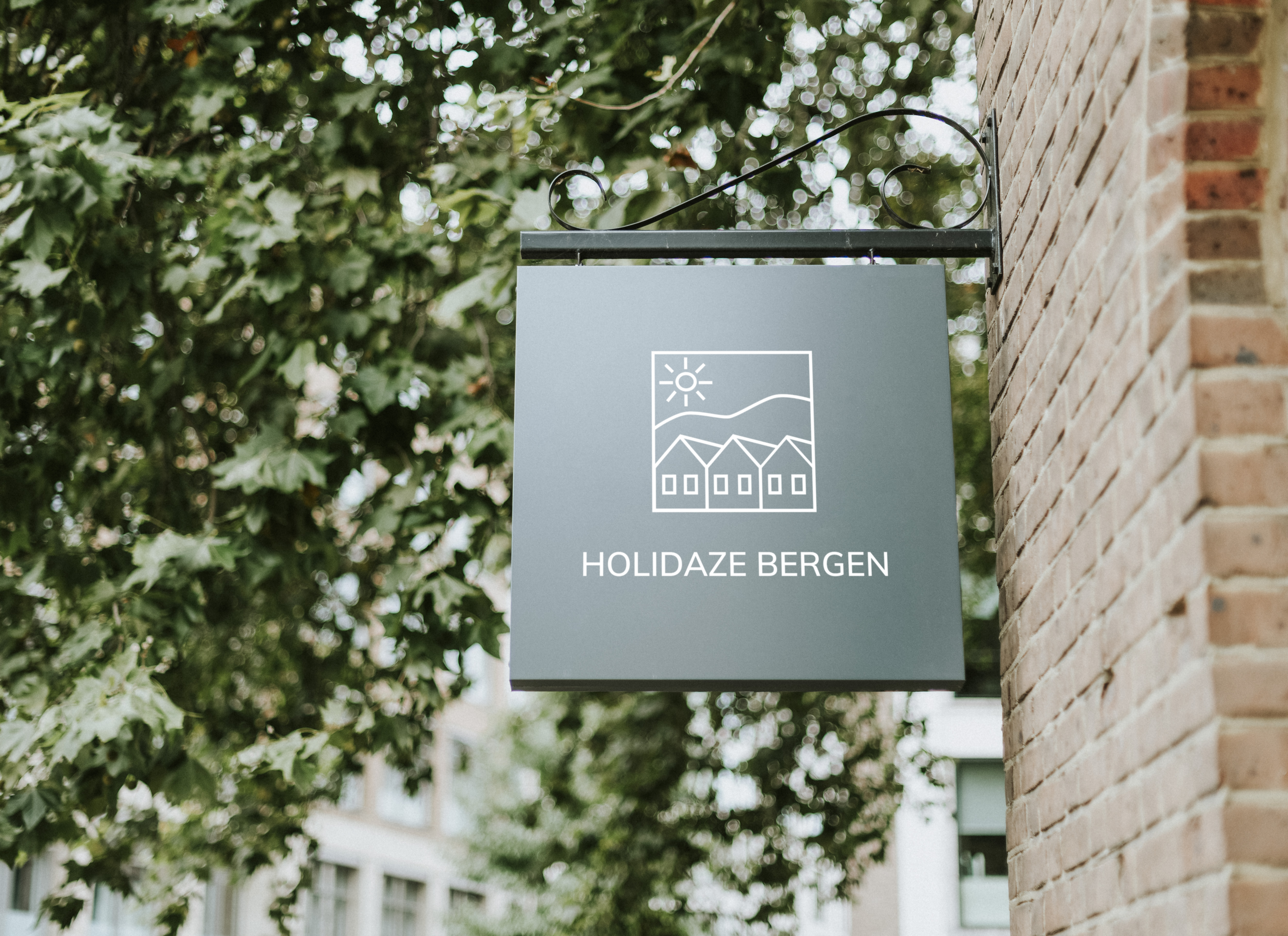 grønt skilt med Holidaze Bergen logo langs murvegg med trær i bakgrunnen